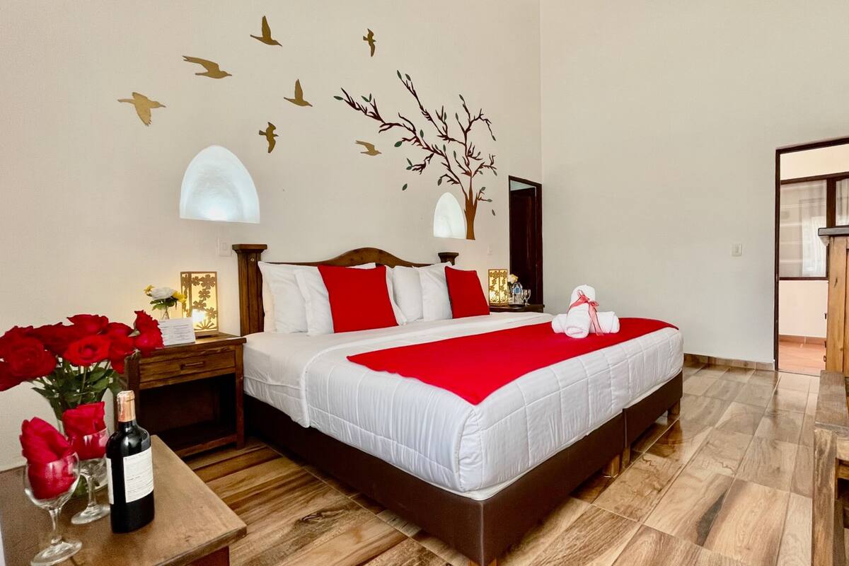 A hotel room with a large bed, wooden headboard, and a red and white bedspread. There is a nightstand with a lamp, a vase of red roses, and a bottle of wine. The room has a wooden floor and a view of the hallway.