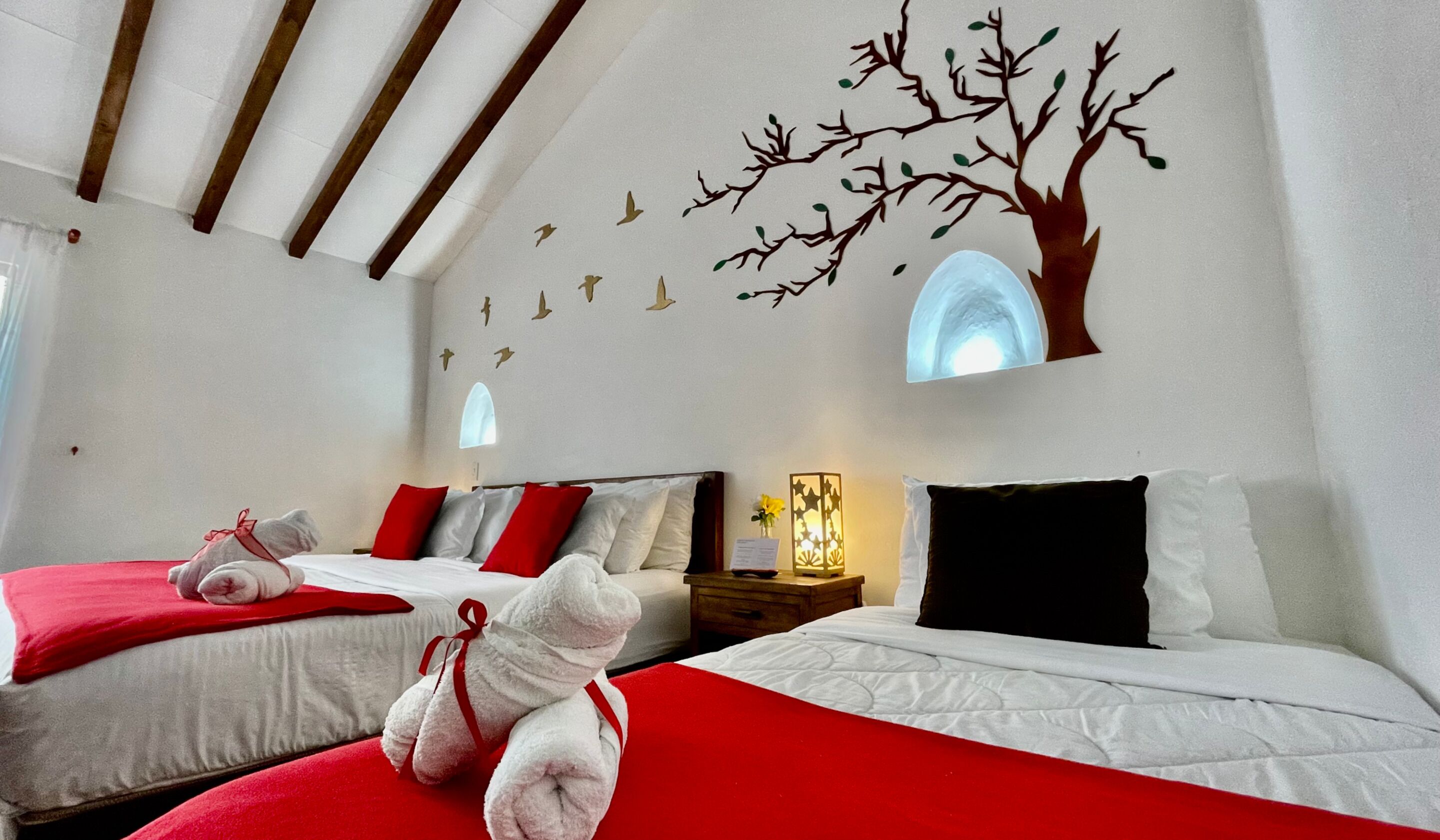 Ein Hotelzimmer mit zwei Betten, roter und weißer Bettwäsche, einem Teddybären, einer Baummalerei und einer beleuchteten Wandlampe.