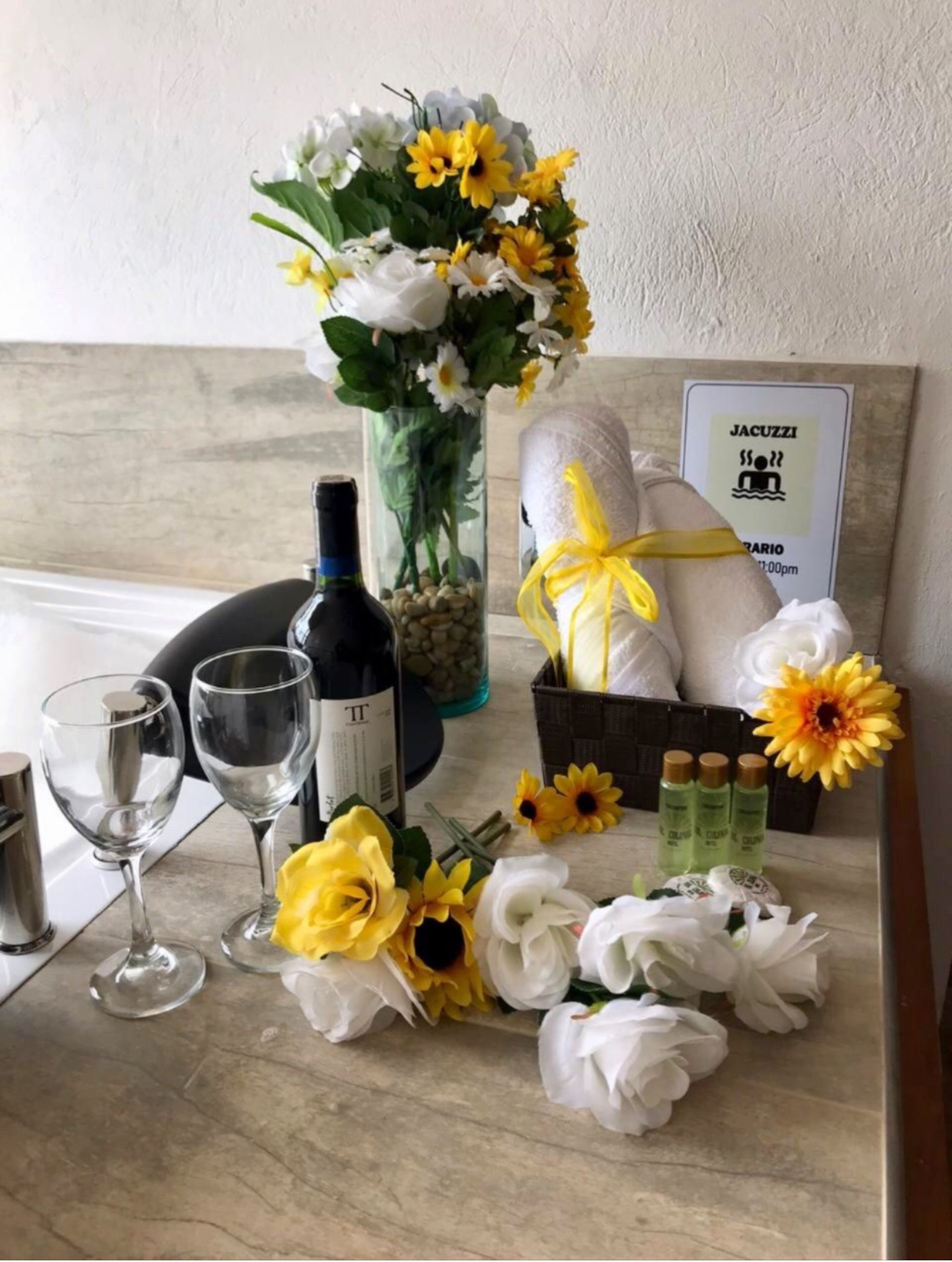 A table with a bottle of wine, two wine glasses, a vase with flowers, a basket of toiletries, and a towel.