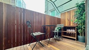 Una terraza de madera con dos sillas y una mesita, un estante de madera con zapatos, y una planta en maceta.