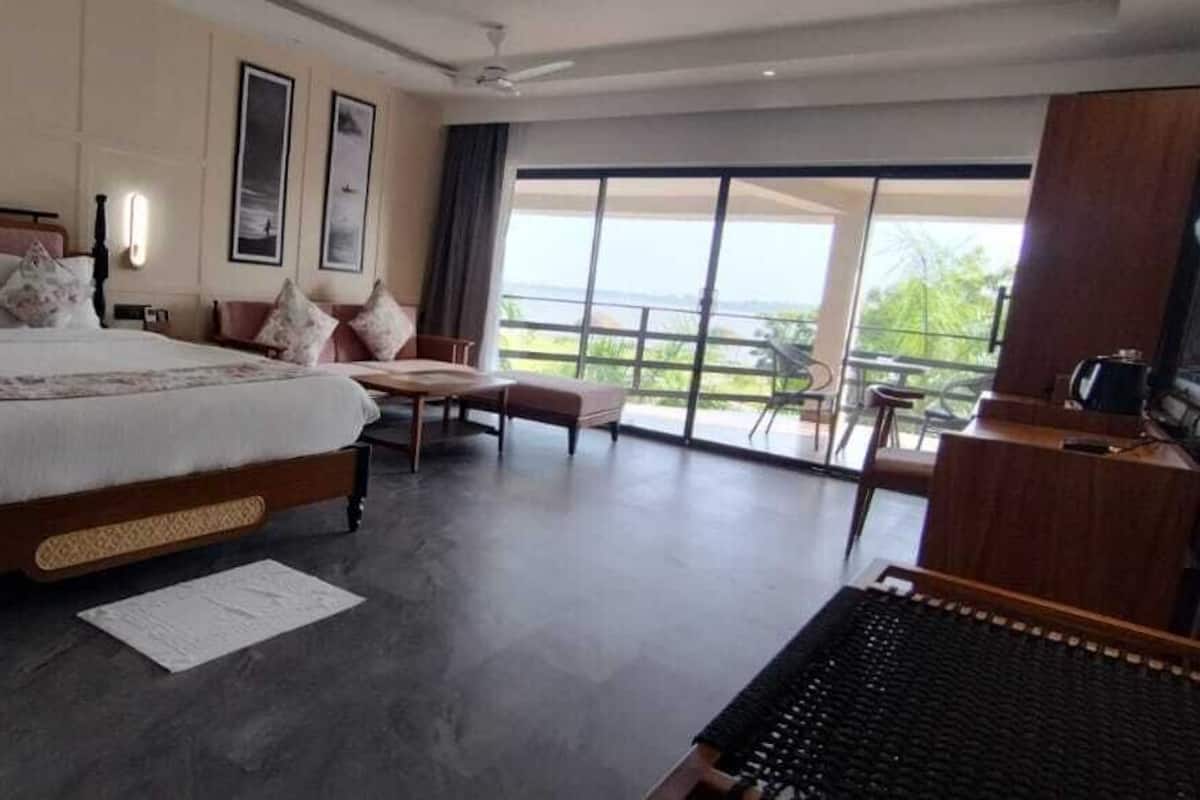 A spacious bedroom with a large bed, a wooden nightstand, a desk, and a chair. There is a balcony with a view of the outdoors.