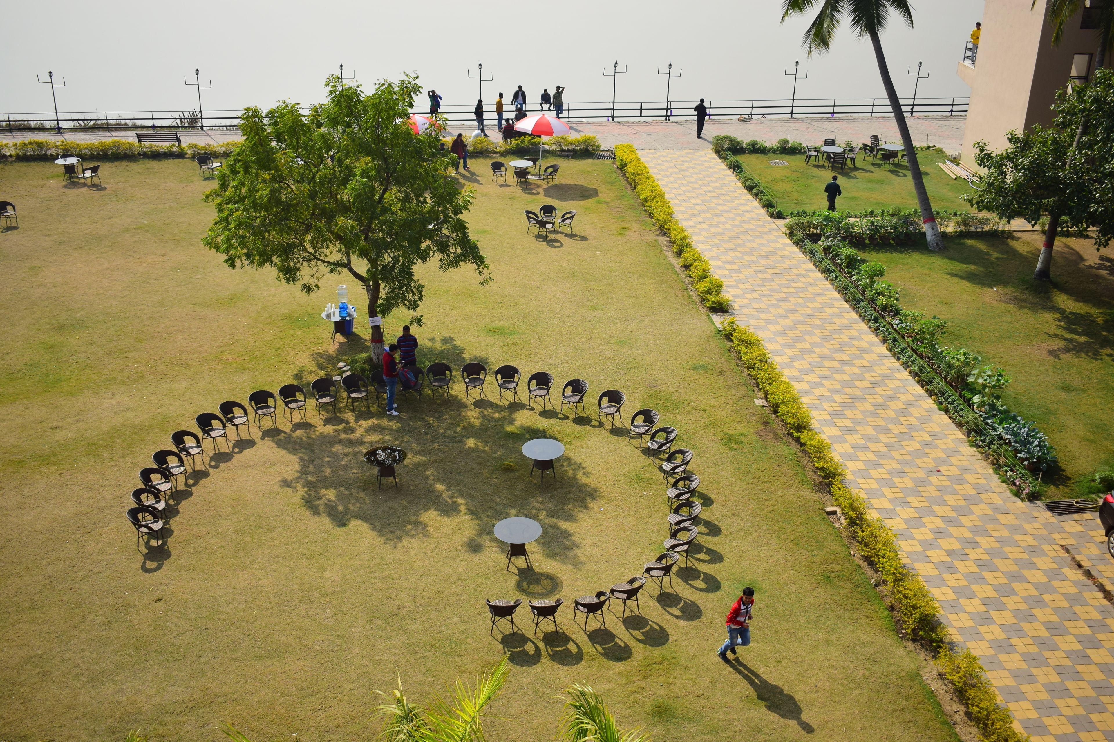 An outdoor seating area with a circular arrangement of tables and chairs, a central tree, and a checkered pathway.