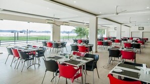 A spacious indoor dining area with tables and chairs, large windows offering a view of a body of water, and ceiling fans.