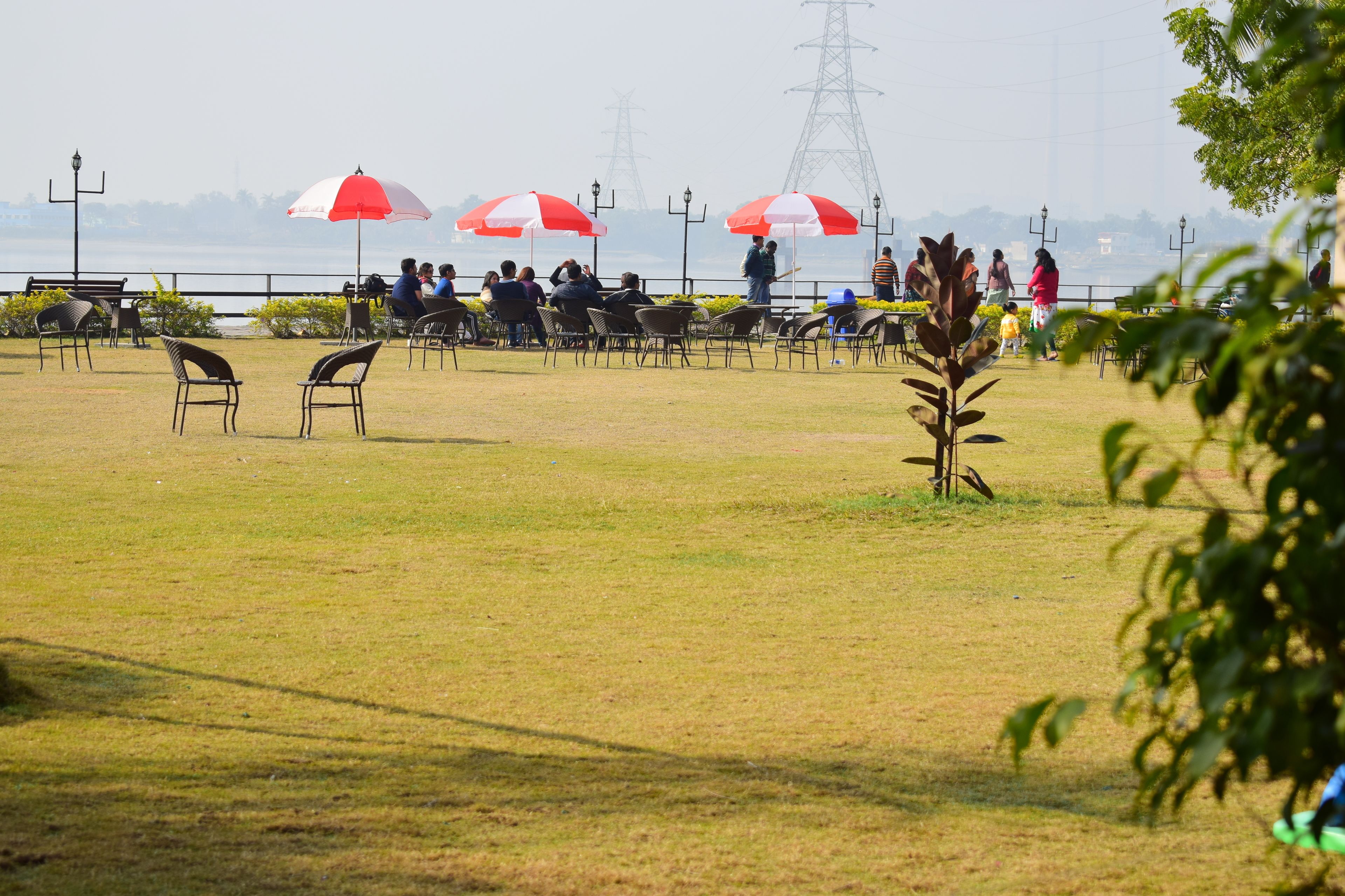 An outdoor seating area with tables and chairs, some under red and white umbrellas, and a view of a body of water with power lines in the background.