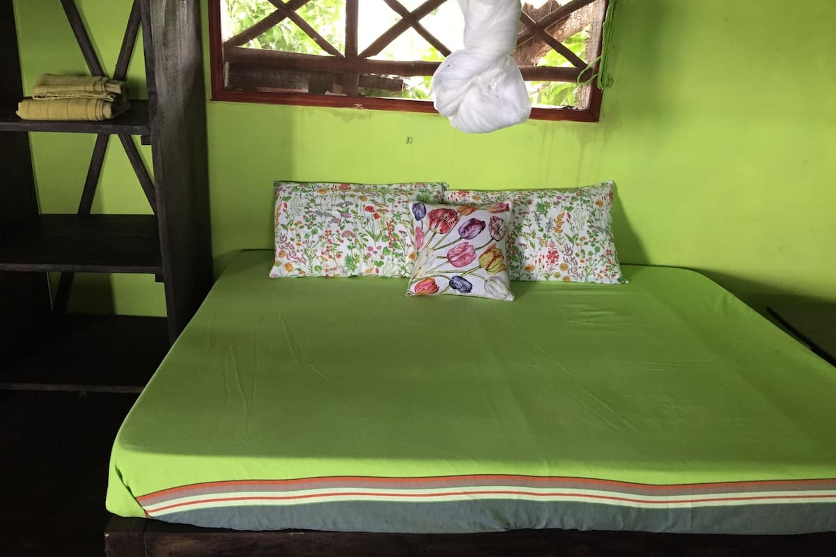 A bed with a green bedspread and floral pillows, a wooden chair, and a window with a white hanging object.
