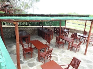 Un espacio de patio al aire libre con varias mesas y sillas de madera, piso de piedra y una estructura de techo verde.