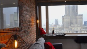 A modern living room with a brick wall, a grey sofa with red cushions, a hanging light fixture, and a city view through large windows. - Panda Apartments Grzybowska-Centrum (Warsaw)