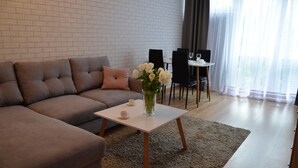 A modern living room with a sectional sofa, a small white table with a vase of flowers, and a dining area with chairs and a table. - Panda Apartments Grzybowska-Centrum (Warsaw)