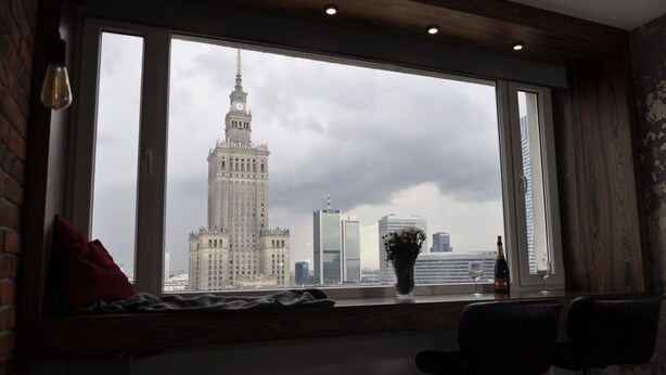 A room with a large window overlooking a cityscape, including a prominent building with a clock tower. - Panda Apartments Grzybowska-Centrum (Warsaw)