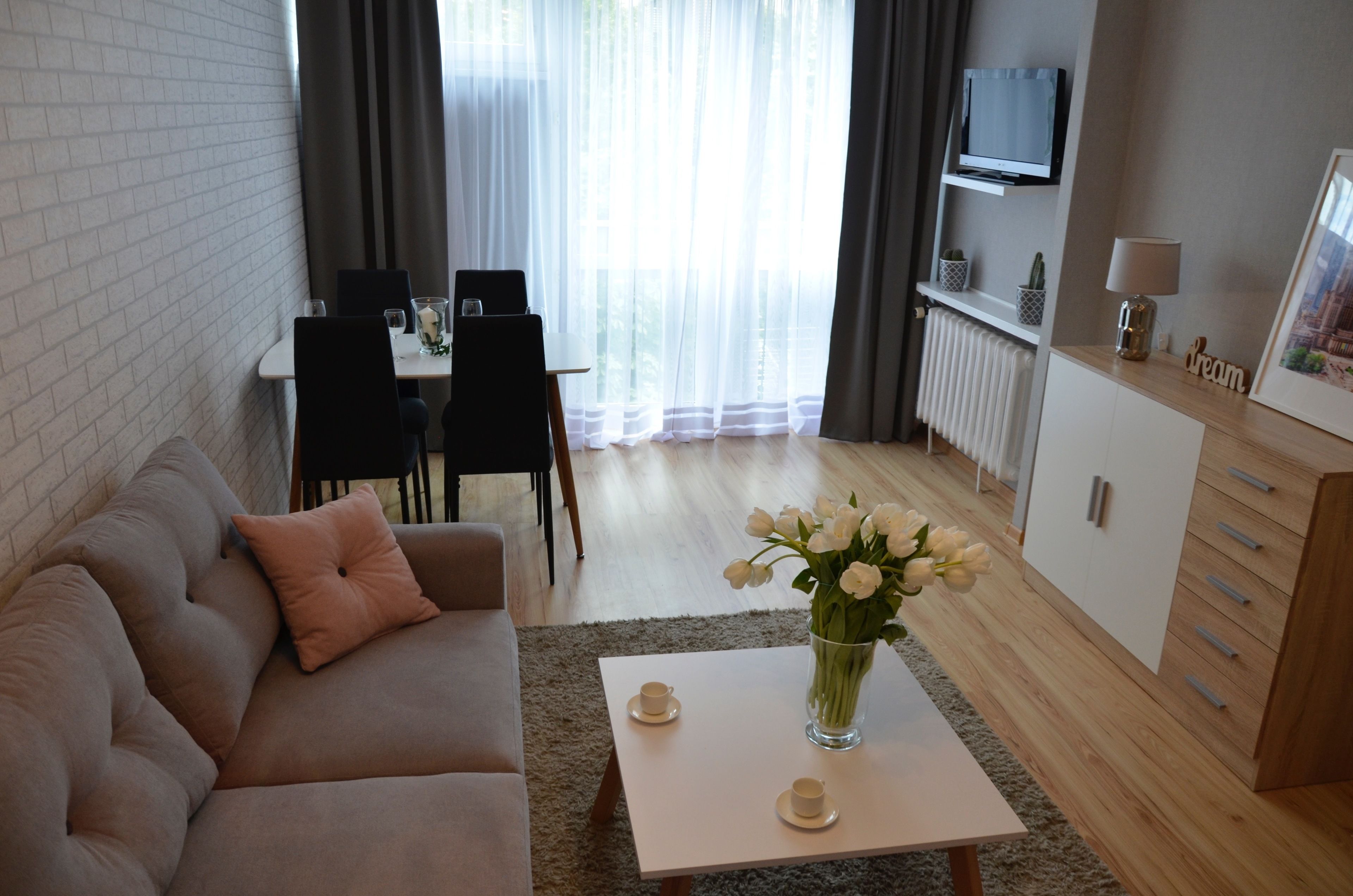 A modern living room with a sofa, a coffee table with a vase of flowers, a dining area with chairs and a table, and a TV mounted on the wall.
