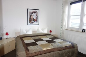 A bedroom with a large bed, a nightstand, a red lamp, a framed picture on the wall, and a window with blinds.