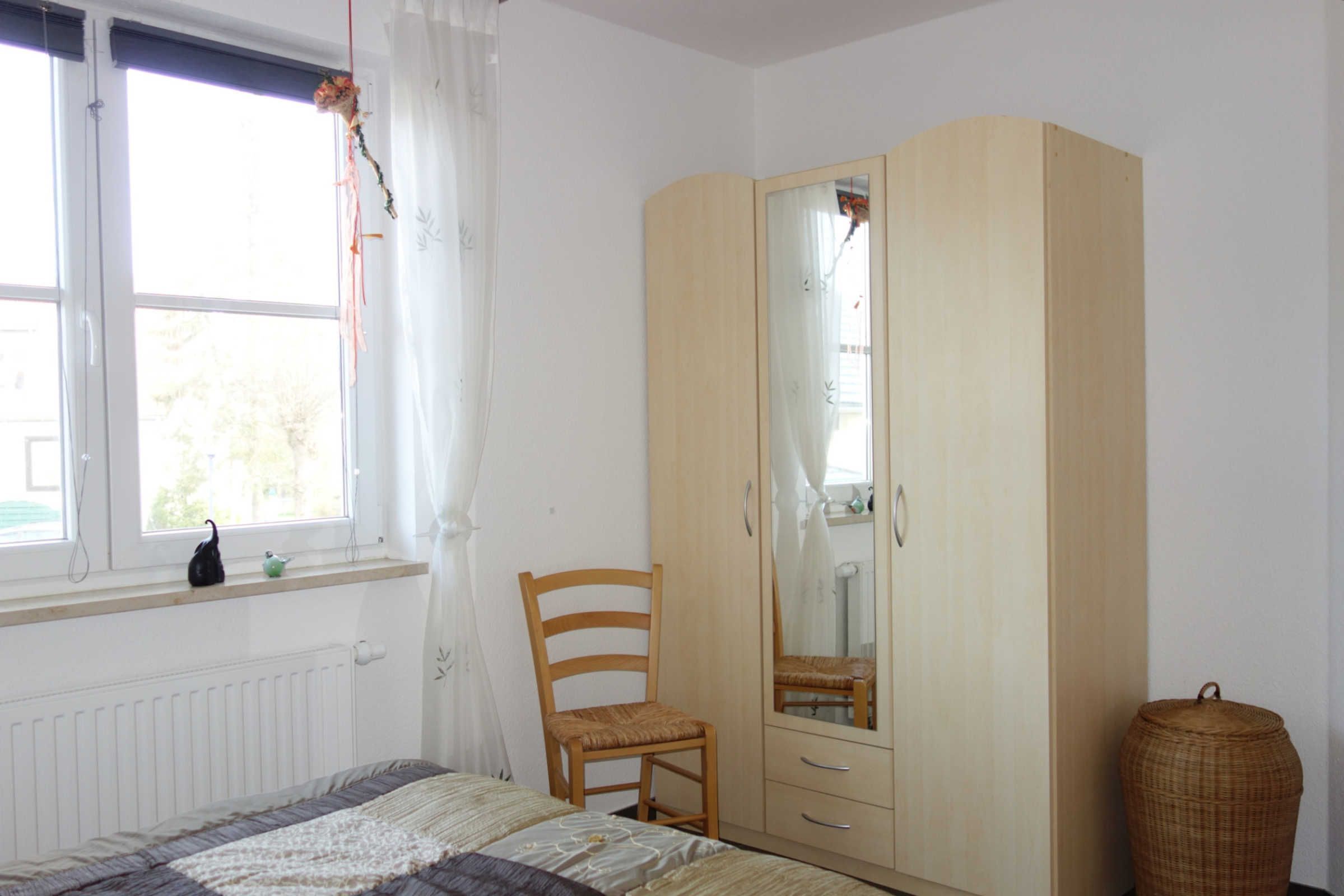 Un dormitorio con una cama, un armario de madera con espejo, una silla, un ventanal con cortinas y un radiador.