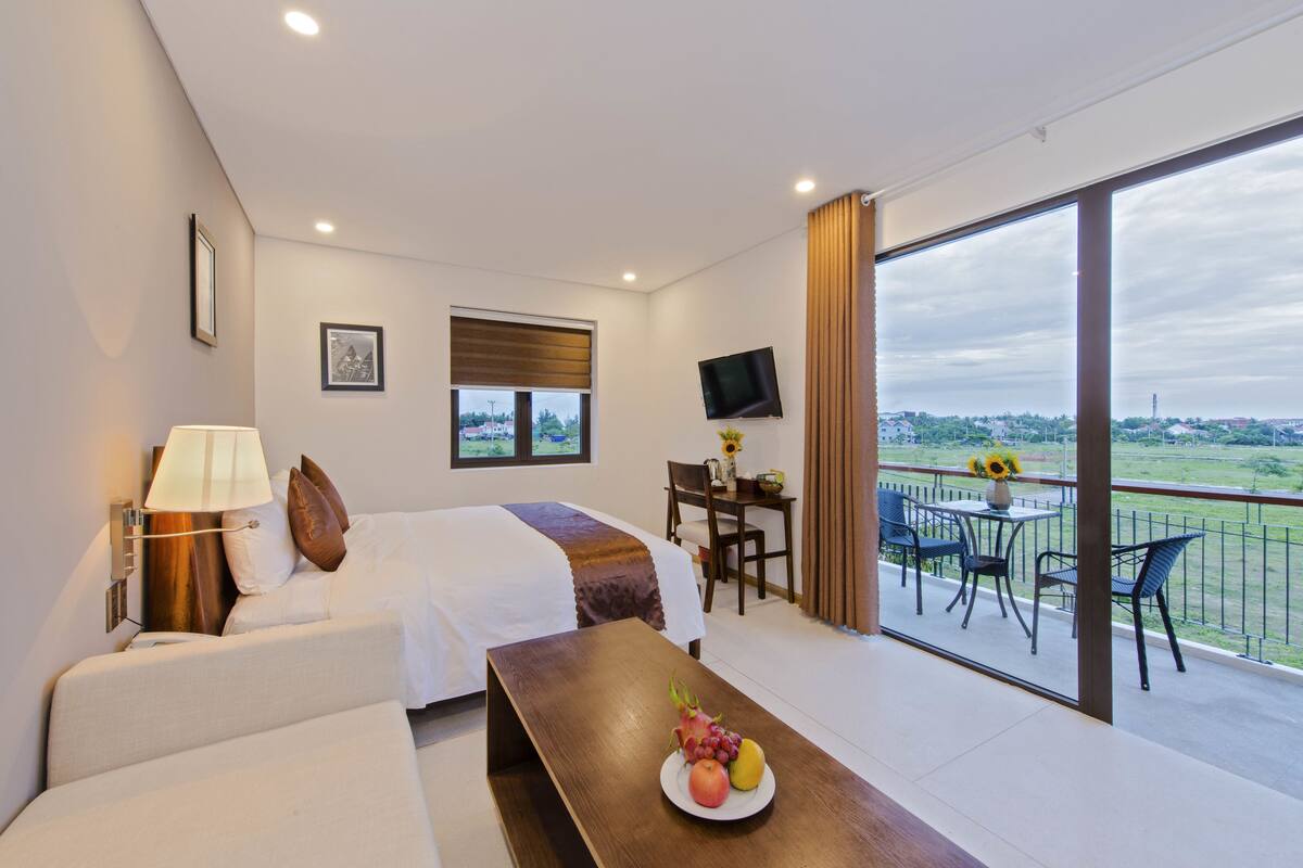 A hotel room with two beds, a desk with a chair, a table with fruit, a TV, and a balcony with a view.