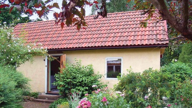 Una pequeña casa de un solo piso con techo de tejas rojas, rodeada de vegetación exuberante y plantas con flores.