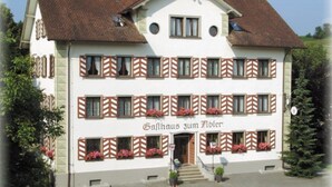 Un bâtiment à deux étages, doté d’une façade blanche et de volets marron. L’enseigne indique : « Gasthof zum Felsen ».