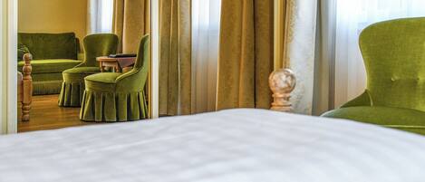 Una habitación de hotel con una cama grande, dos sillones verdes, una mesita de madera y un televisor en la pared.