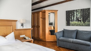Une chambre d’hôtel avec une armoire en bois, un canapé gris à pois, un lit, une table de chevet avec une lampe et un tableau sur le thème de la forêt accroché au mur.