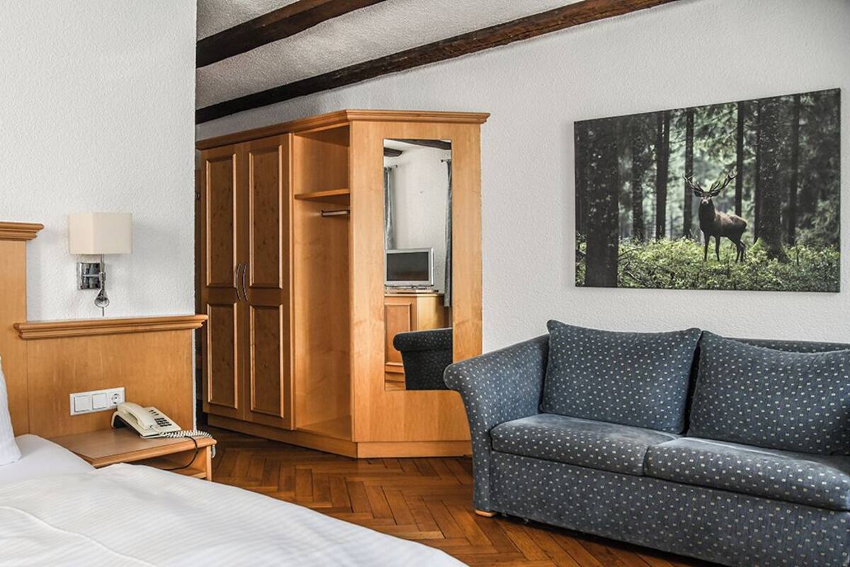 Hotelkamer met een houten garderobe, een grijze stippenbank, een bed, een nachtkastje met een lamp, en een schilderij met een boslandschap op de muur.