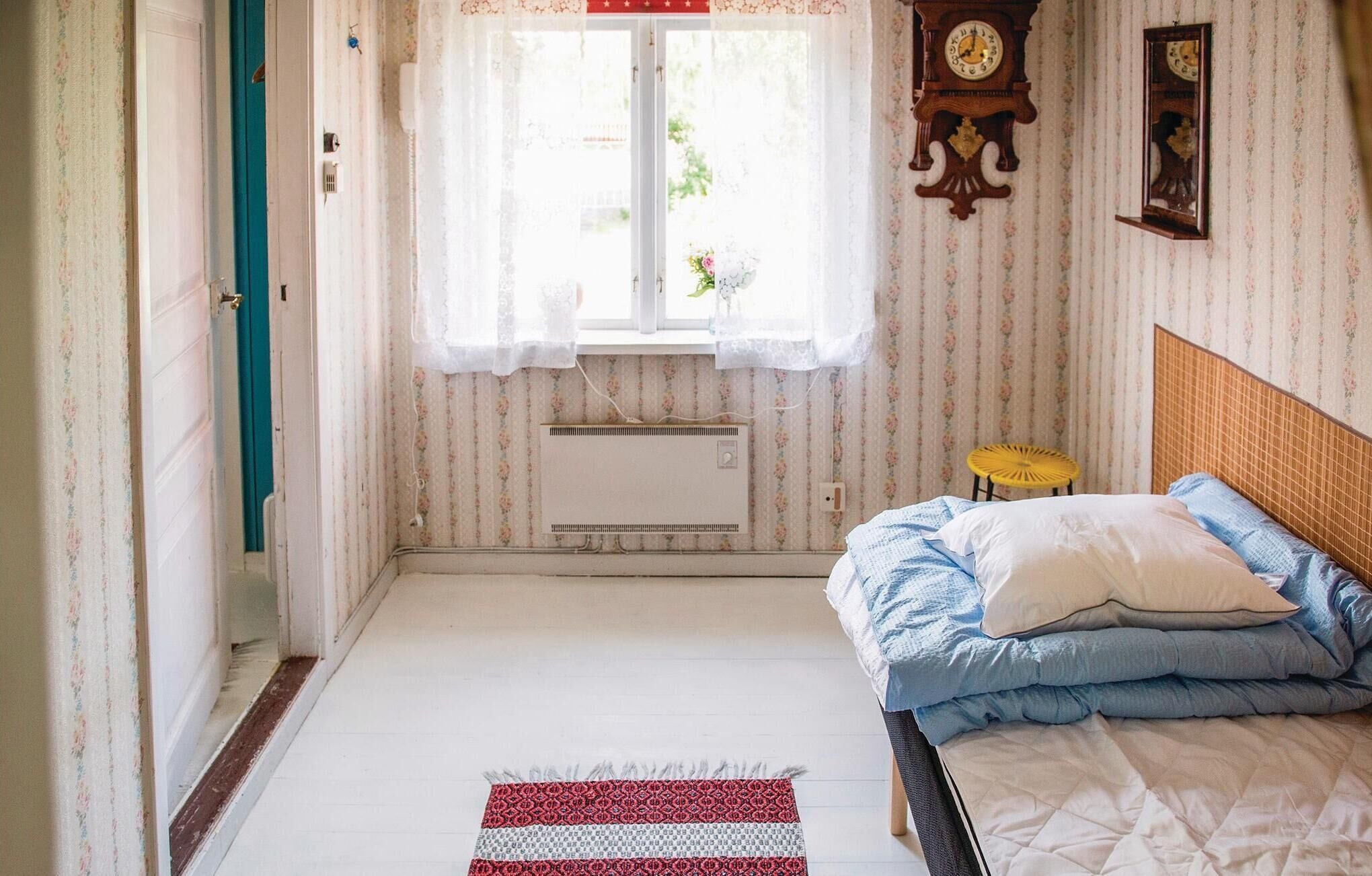 A bedroom with a bed, a window with lace curtains, a radiator, and a clock on the wall.