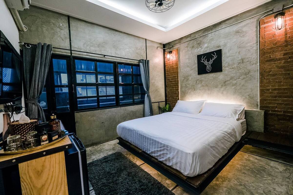Quarto moderno com cama, escrivaninha com máquina de café e uma janela grande com cortinas.