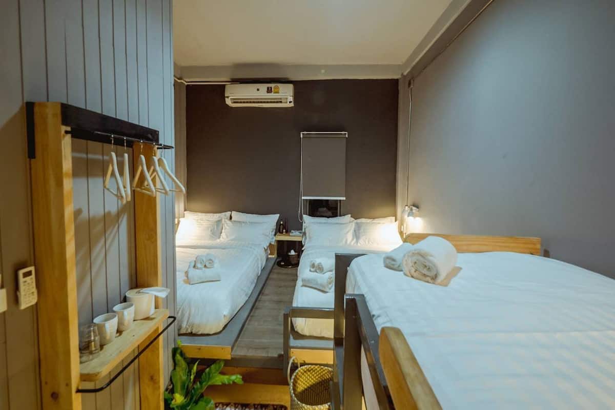 Quarto de hotel com três camas, um guarda-roupa de madeira com roupas penduradas, uma mesa pequena com chávenas e um ar condicionado fixado na parede.