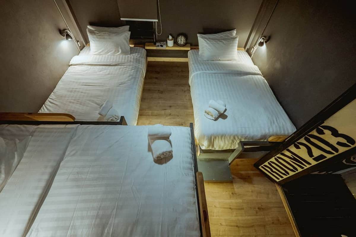 Quarto com três camas alinhadas, cada uma com almofada e uma toalha enrolada. Há uma pequena prateleira com um relógio e uma caixa de lenços. O quarto possui piso de madeira e uma porta visível com um sinal de número.