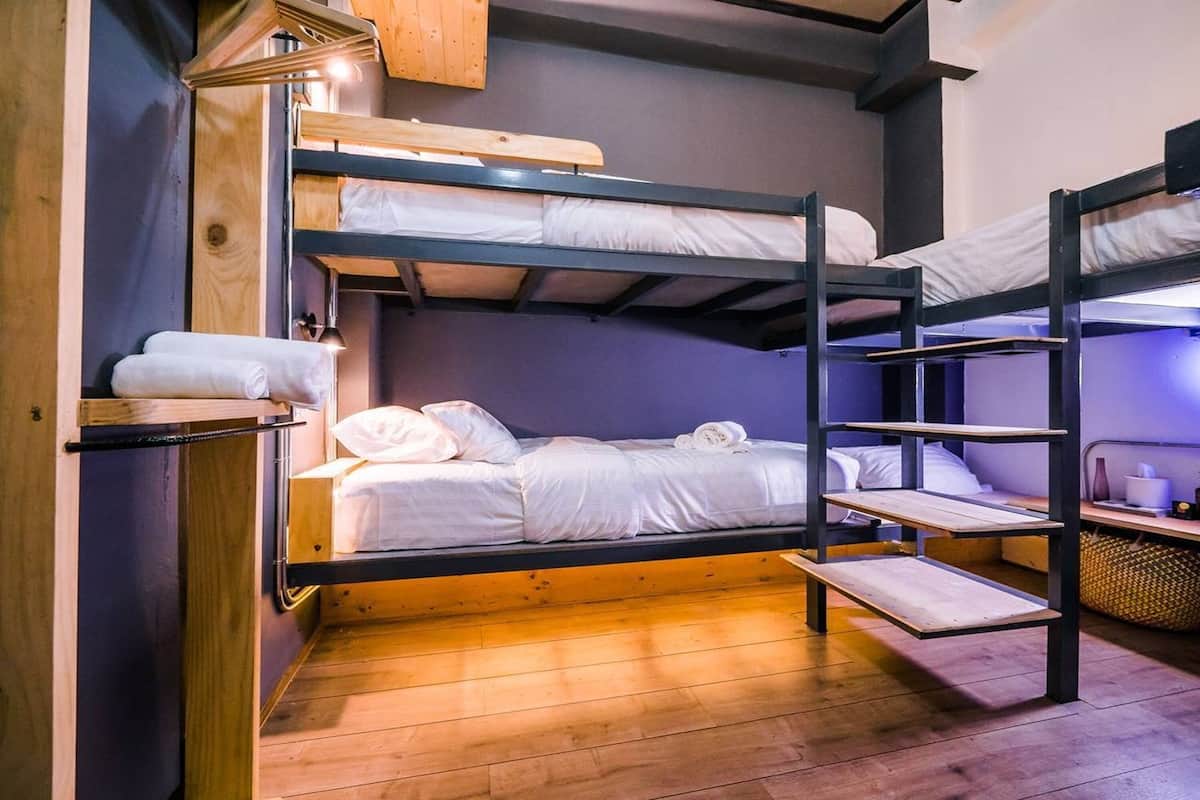 Quarto com beliches, piso de madeira, prateleira com toalhas e uma luminária.