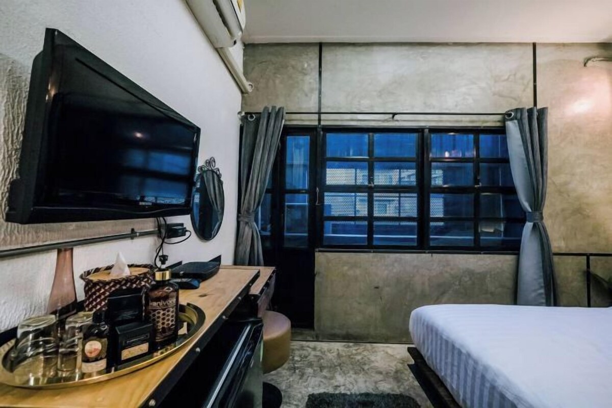 A hotel room with a bed, a TV mounted on the wall, a bedside table with a tray of toiletries, and a window with curtains.