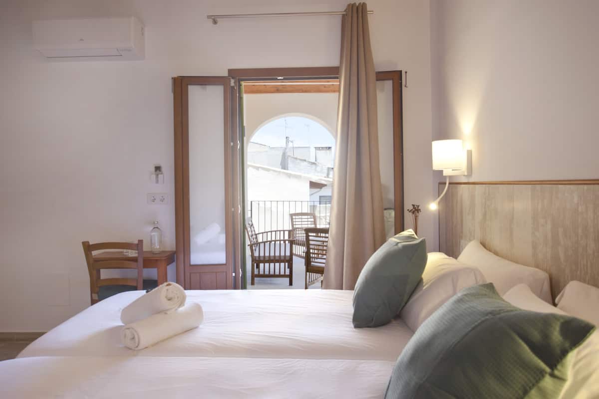 Habitación de hotel con cama, almohadas, una silla, una mesa y una puerta abierta que da a un balcón.