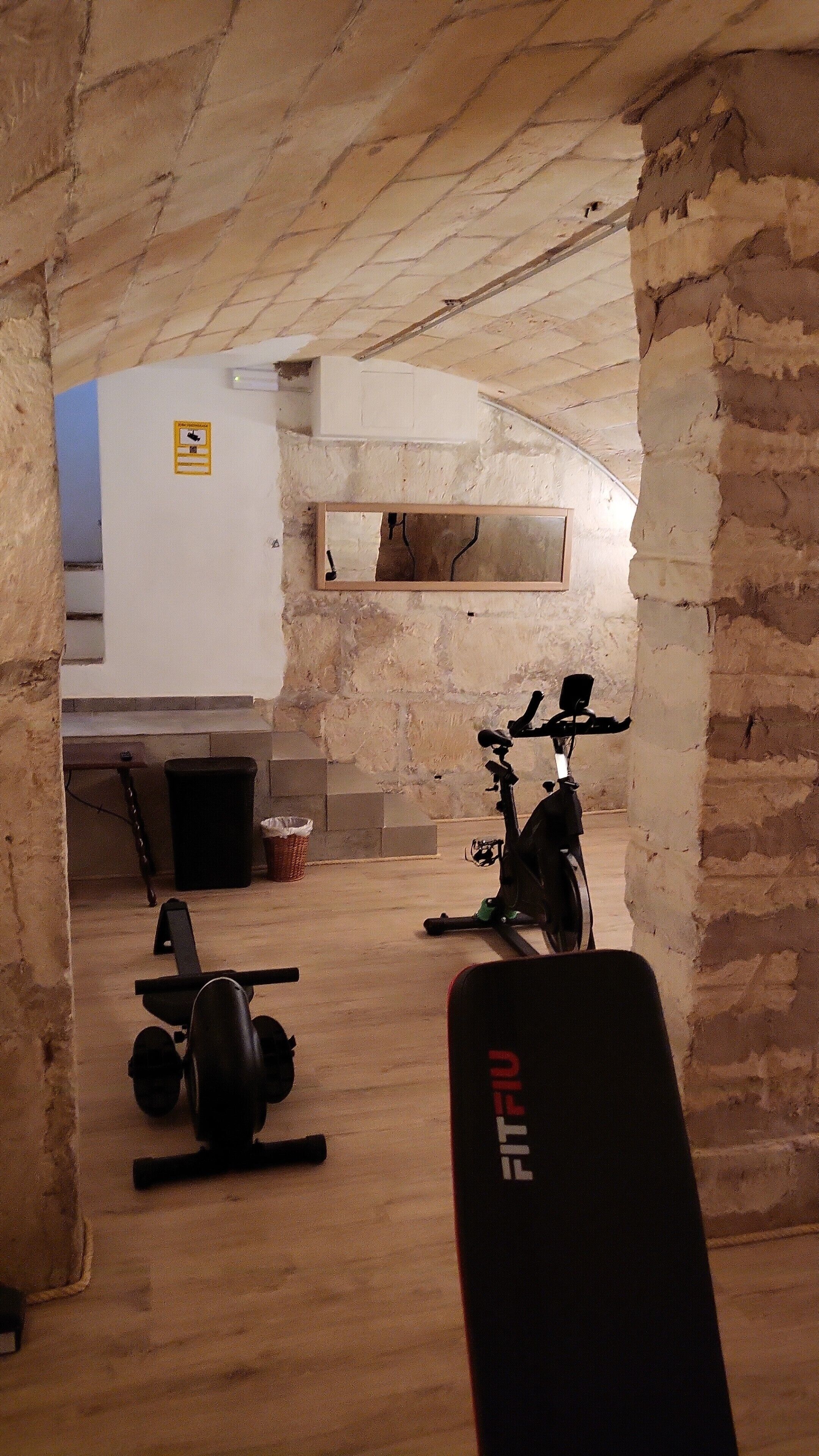 An underground gym with exercise equipment, a white wall with a sign, and exposed brick walls.
