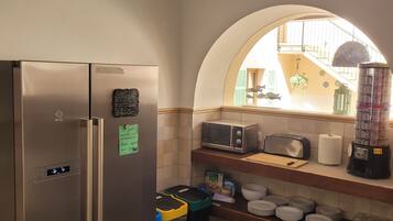 A compact kitchen with a stainless steel refrigerator, a microwave, and a blender on the counter. There are trash bins and a small shelf with dishes.