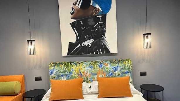 A modern hotel room with a large bed, two orange pillows, a colorful headboard, and two pendant lights.