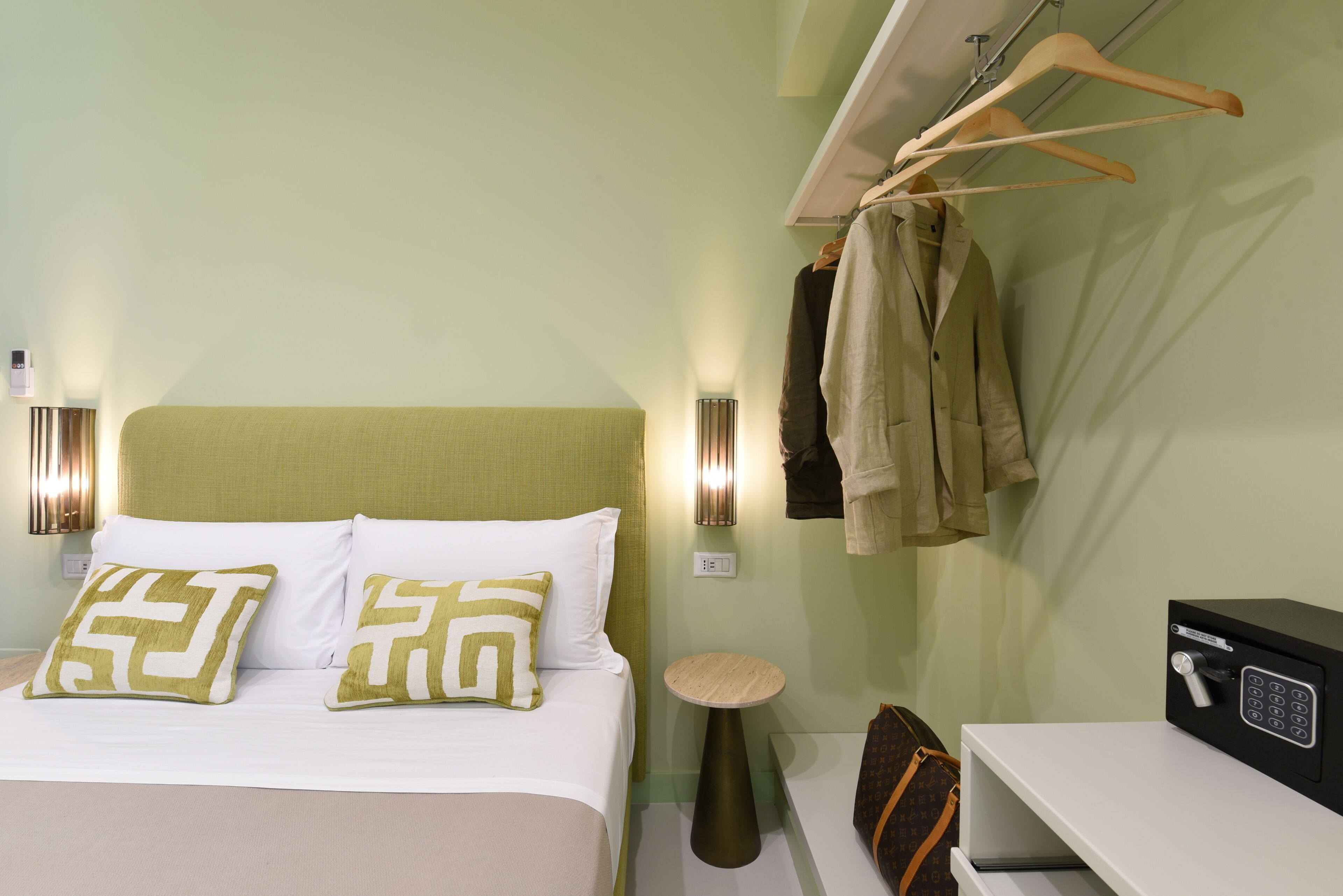 A neatly arranged hotel room with a bed, bedside tables, a lamp, a safe, and a hanging coat rack.