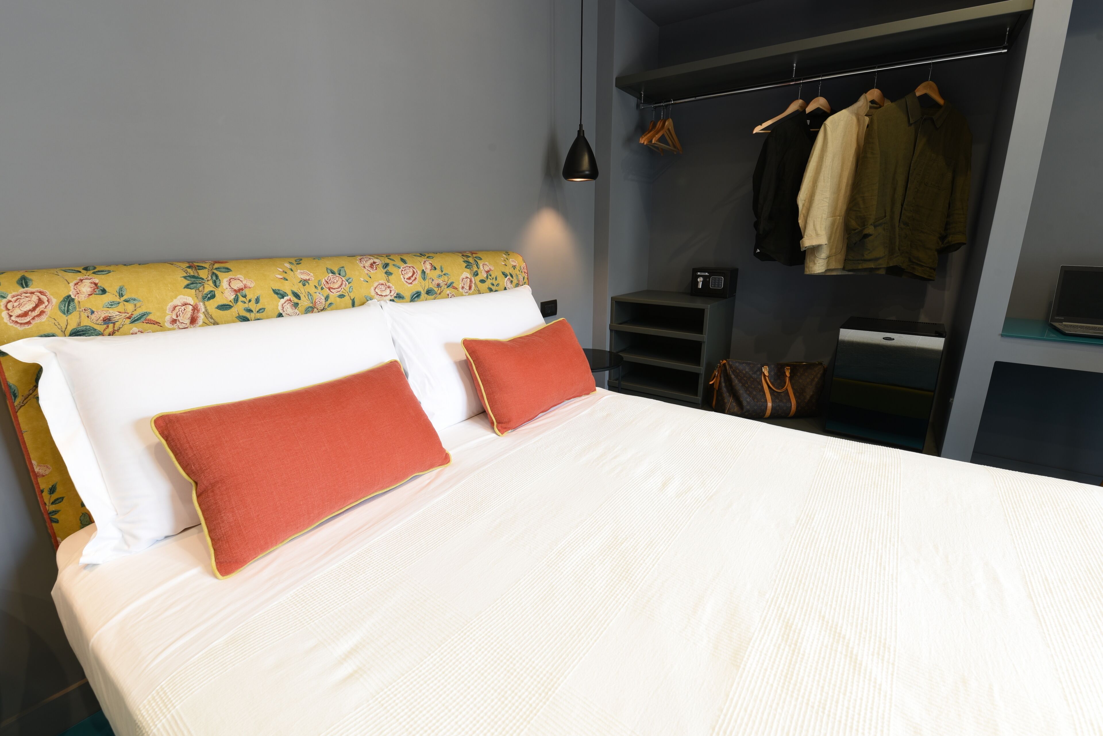 Una cama bien hecha con ropa blanca y dos almohadas rojas, una mesita de noche pequeña, una lámpara colgante y un perchero con varias prendas.