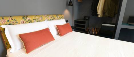 Una cama bien hecha con ropa blanca y dos almohadas rojas, una mesita de noche pequeña, una lámpara colgante y un perchero con varias prendas.