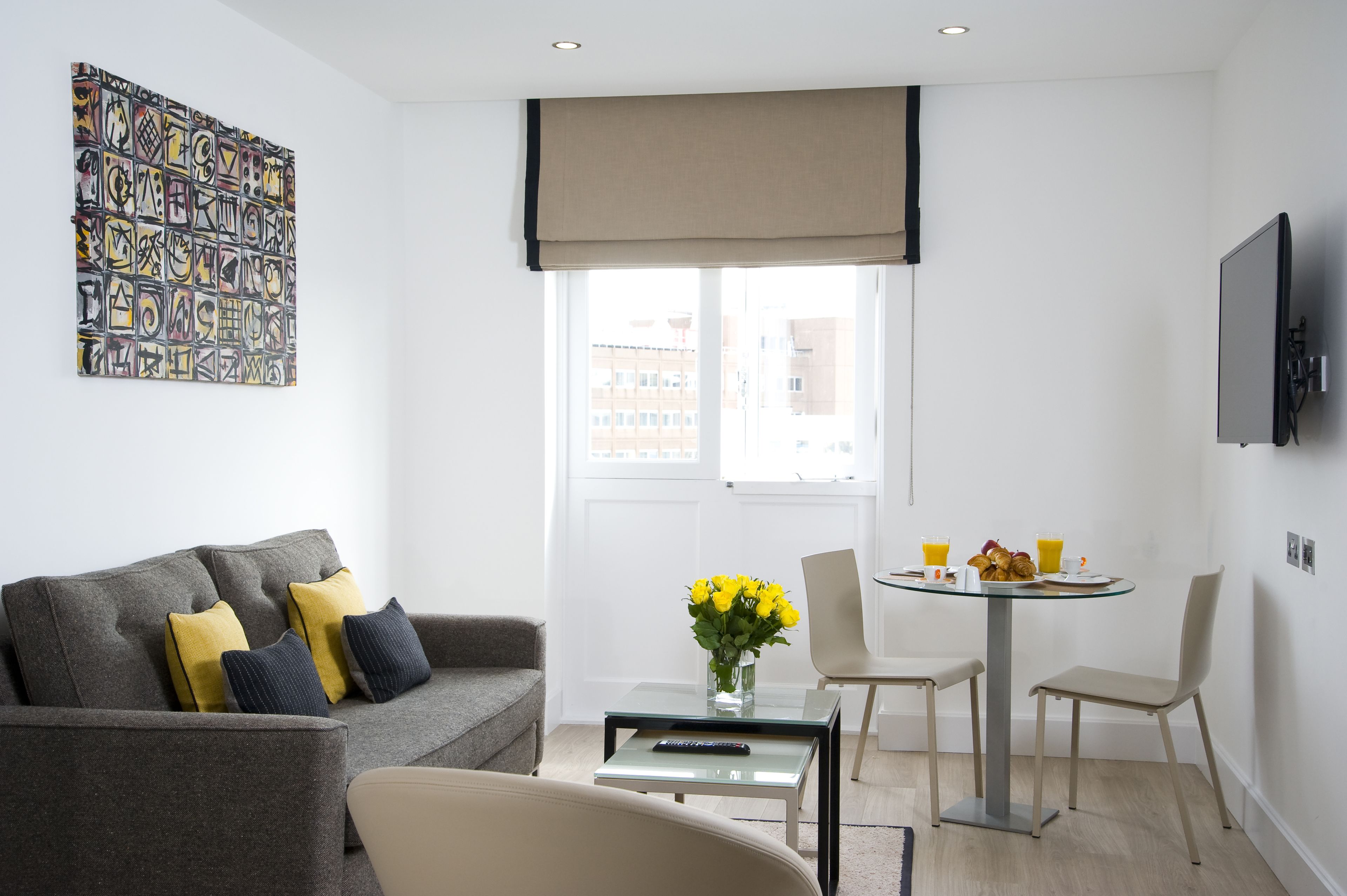 A modern living room with a grey sofa, yellow cushions, a glass coffee table, and a dining area with a small round table and chairs.