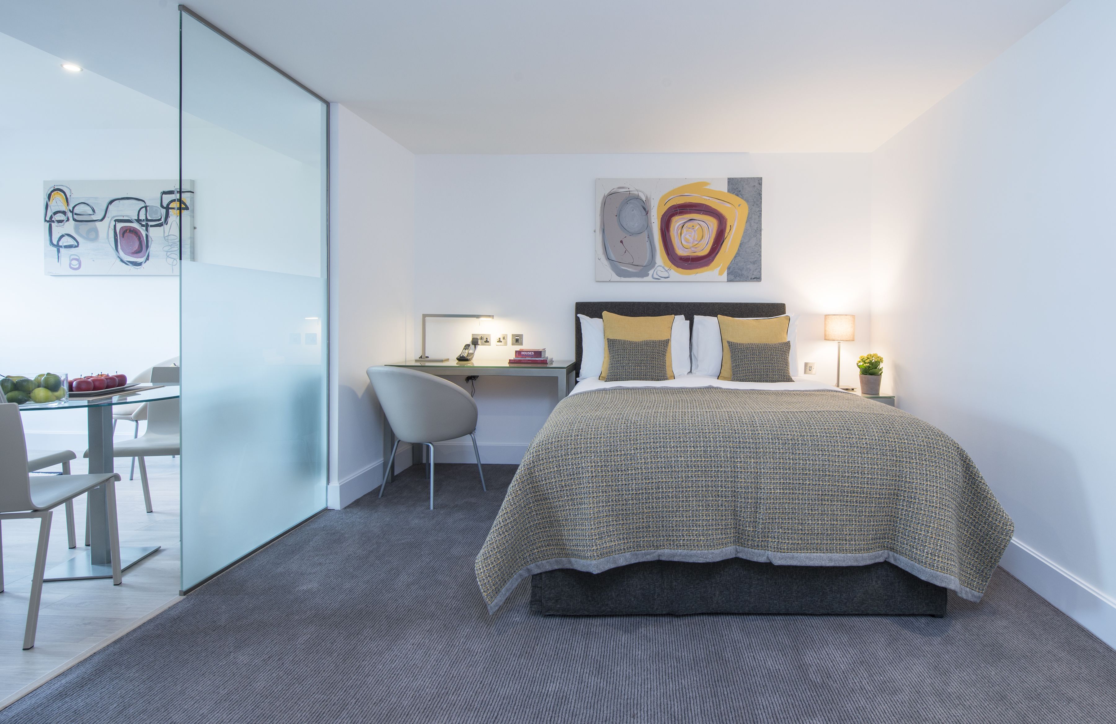 A modern hotel room with a glass partition separating the bedroom from the dining area, a neatly made bed with a patterned bedspread, and a desk with a lamp and a small potted plant.