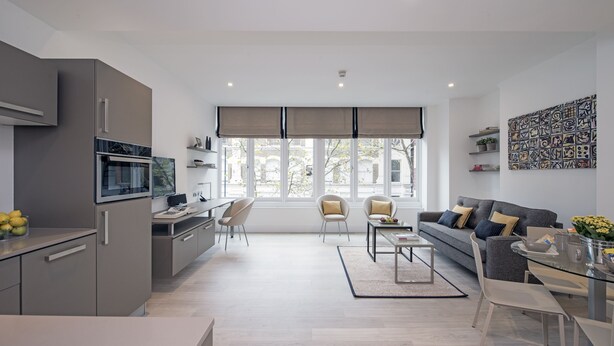 A modern living room with a grey sofa, a coffee table, and a dining area with a glass table. There is a kitchen area with a fridge, oven, and microwave. The room has large windows with blinds and a view of the outside.