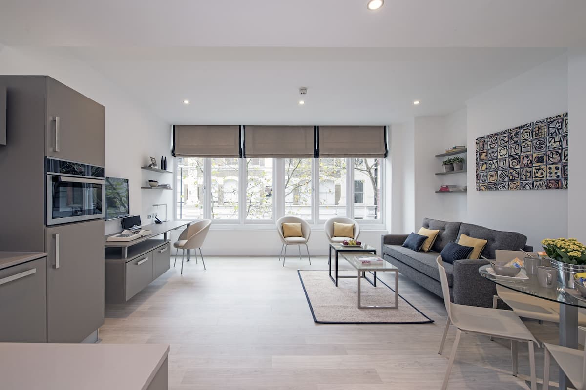 Una sala moderna con un sofá gris, una mesa de centro y un comedor con una mesa de vidrio. Hay una cocina con refrigerador, horno y microondas. La habitación tiene ventanas grandes con persianas y vistas al exterior.
