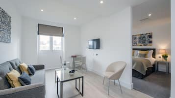 Una habitación de hotel moderna con un sofá gris, una mesita, un televisor, una cama con edredón gris y amarillo, y una ventana con vistas a edificios.