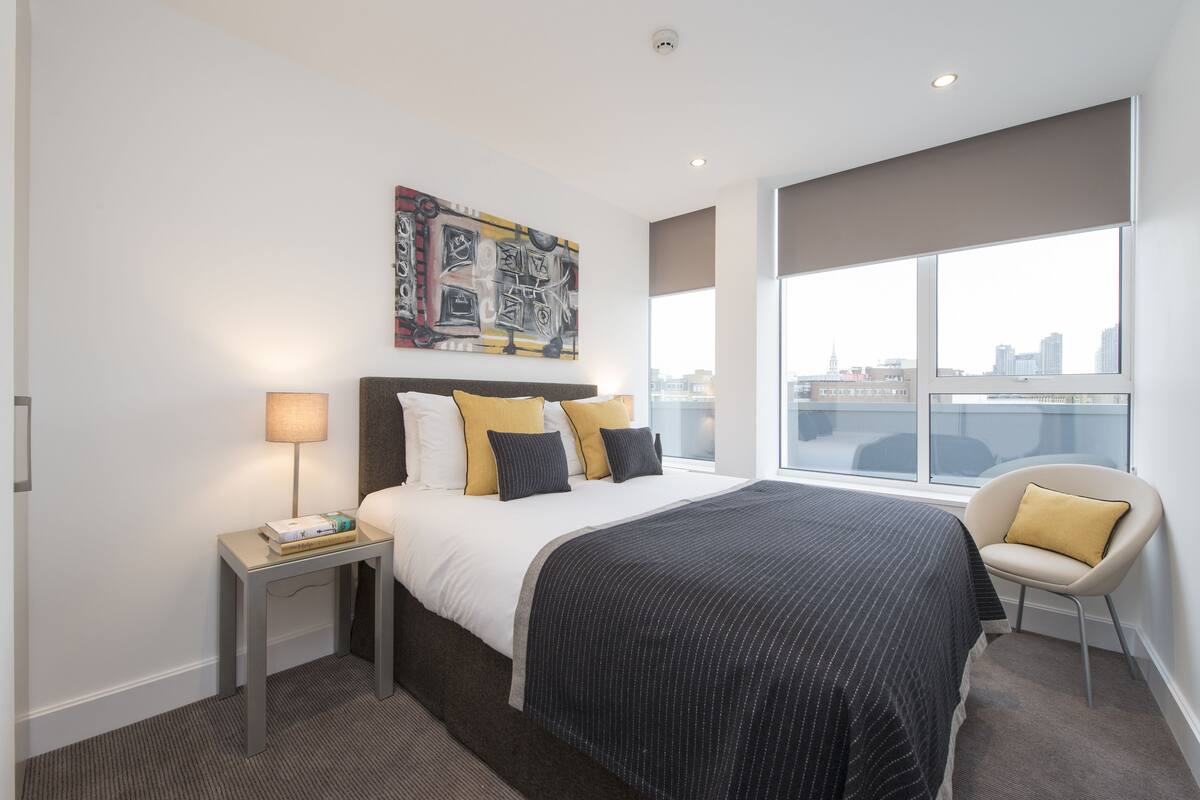 Una habitación de hotel moderna con una cama grande, una mesita de noche, una silla, vista a la ciudad y un cuadro en la pared.