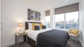 A modern hotel room with a large bed, a nightstand, a chair, a city view, and a painting on the wall.