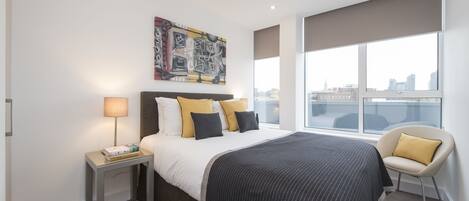 A modern hotel room with a large bed, a nightstand, a chair, a city view, and a painting on the wall.