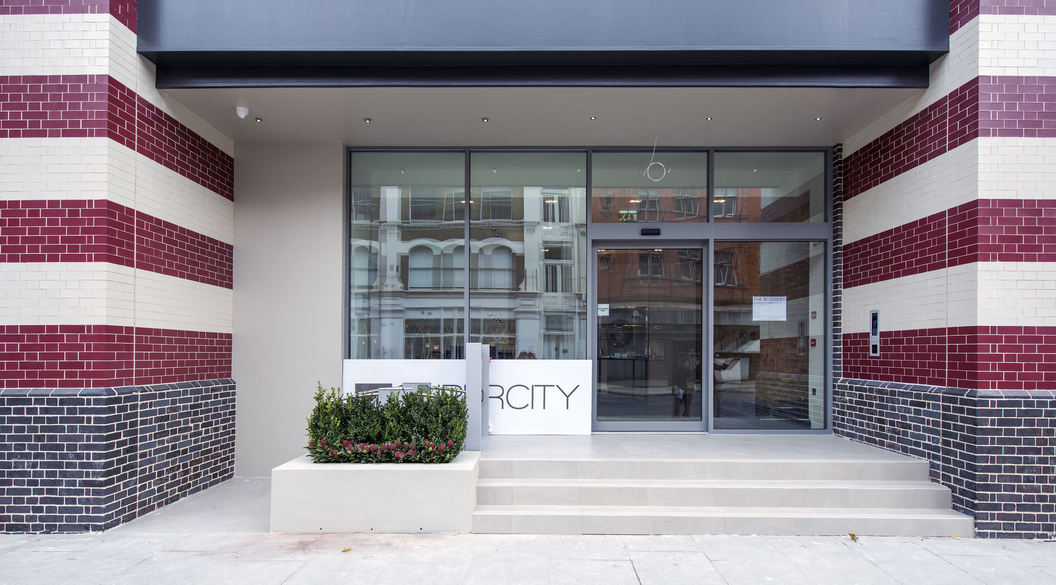A modern building entrance with a glass door, a sign reading "The Rosebery," and a small garden area with plants.