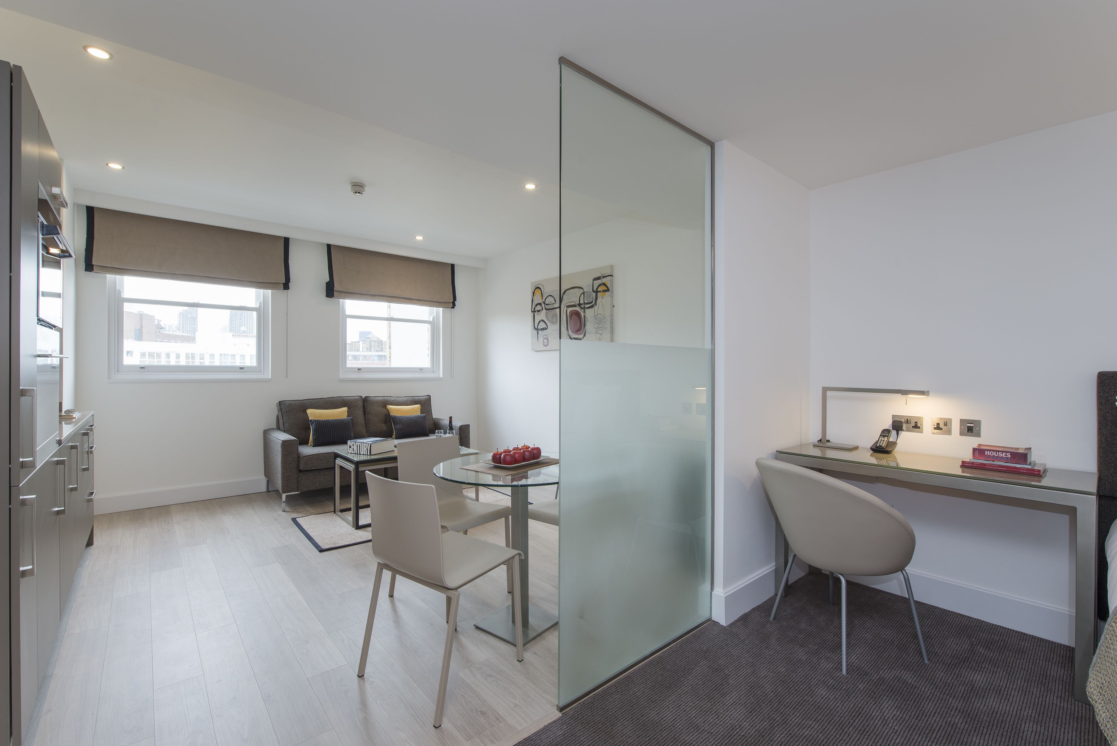 A modern, well-lit room with a glass partition separating a dining area and a workspace. There is a sofa with cushions, a dining table with chairs, and a desk with a chair and a lamp.