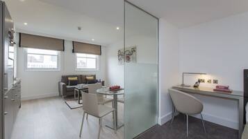 A modern, well-lit room with a glass partition separating a dining area and a workspace. There is a sofa with cushions, a dining table with chairs, and a desk with a chair and a lamp.