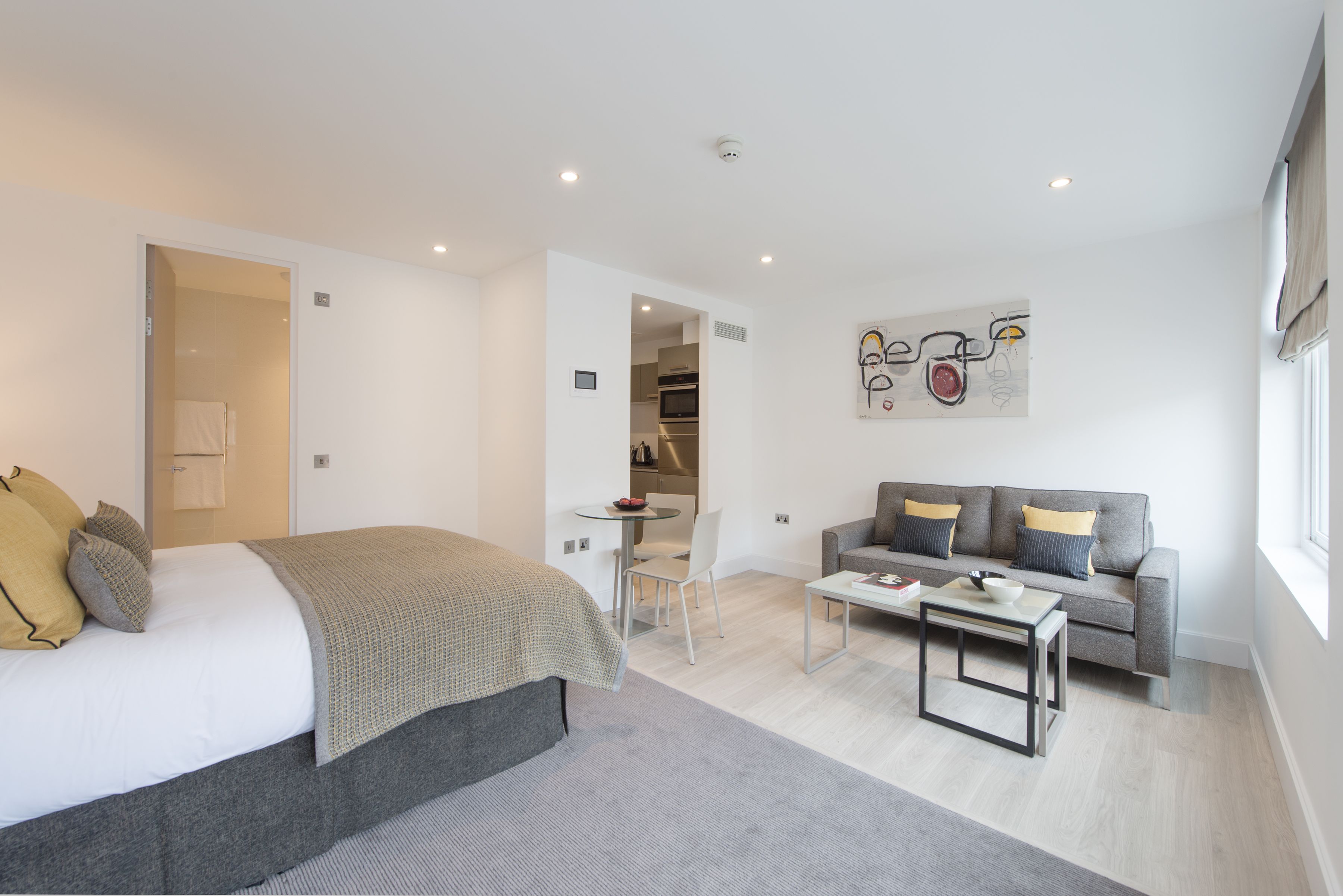 A modern hotel room with a bed, a gray sofa, a small table, and a kitchenette in the background.