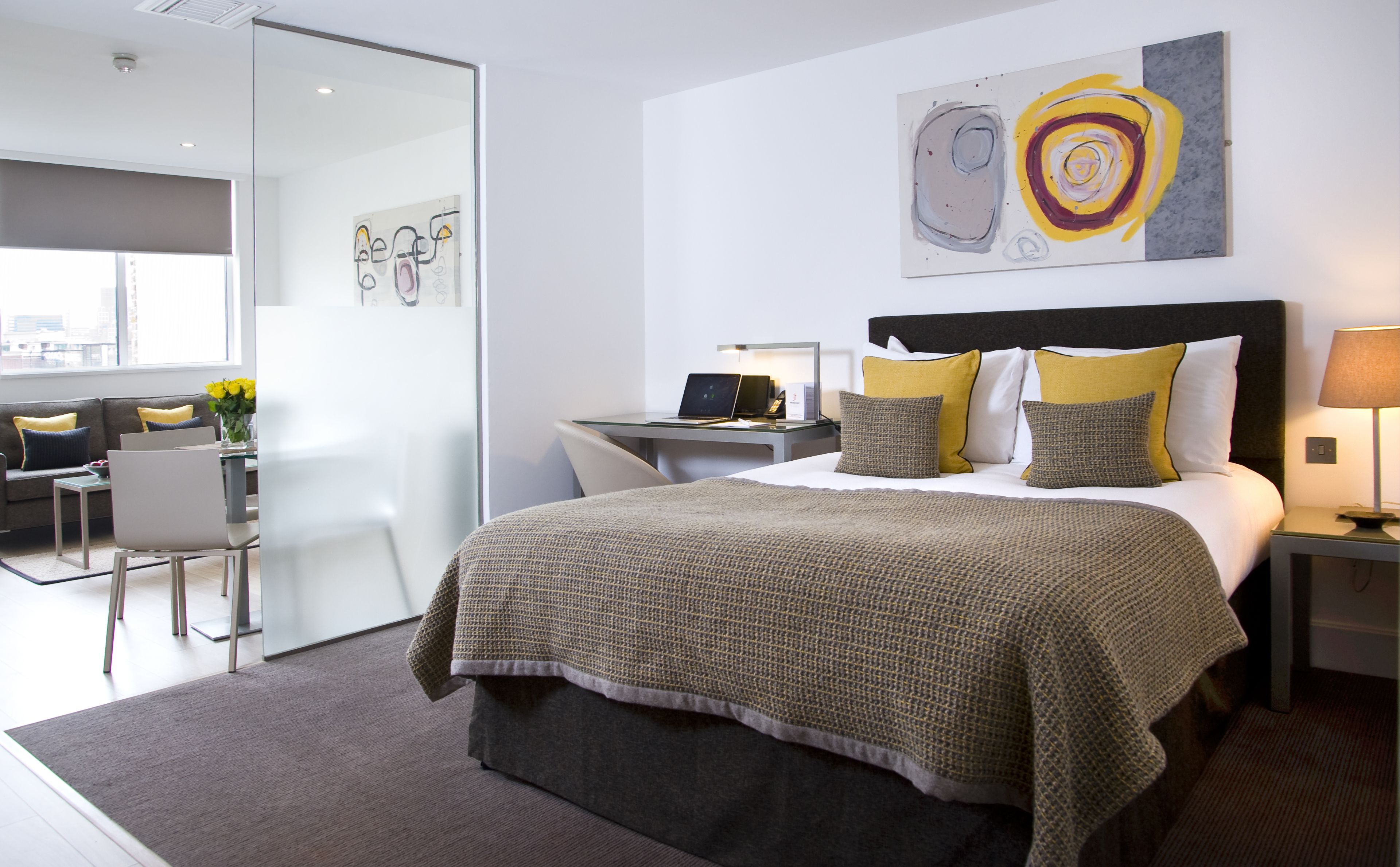 Une chambre d’hôtel moderne avec un grand lit, un bureau avec un ordinateur portable, une chaise et une cloison en verre.