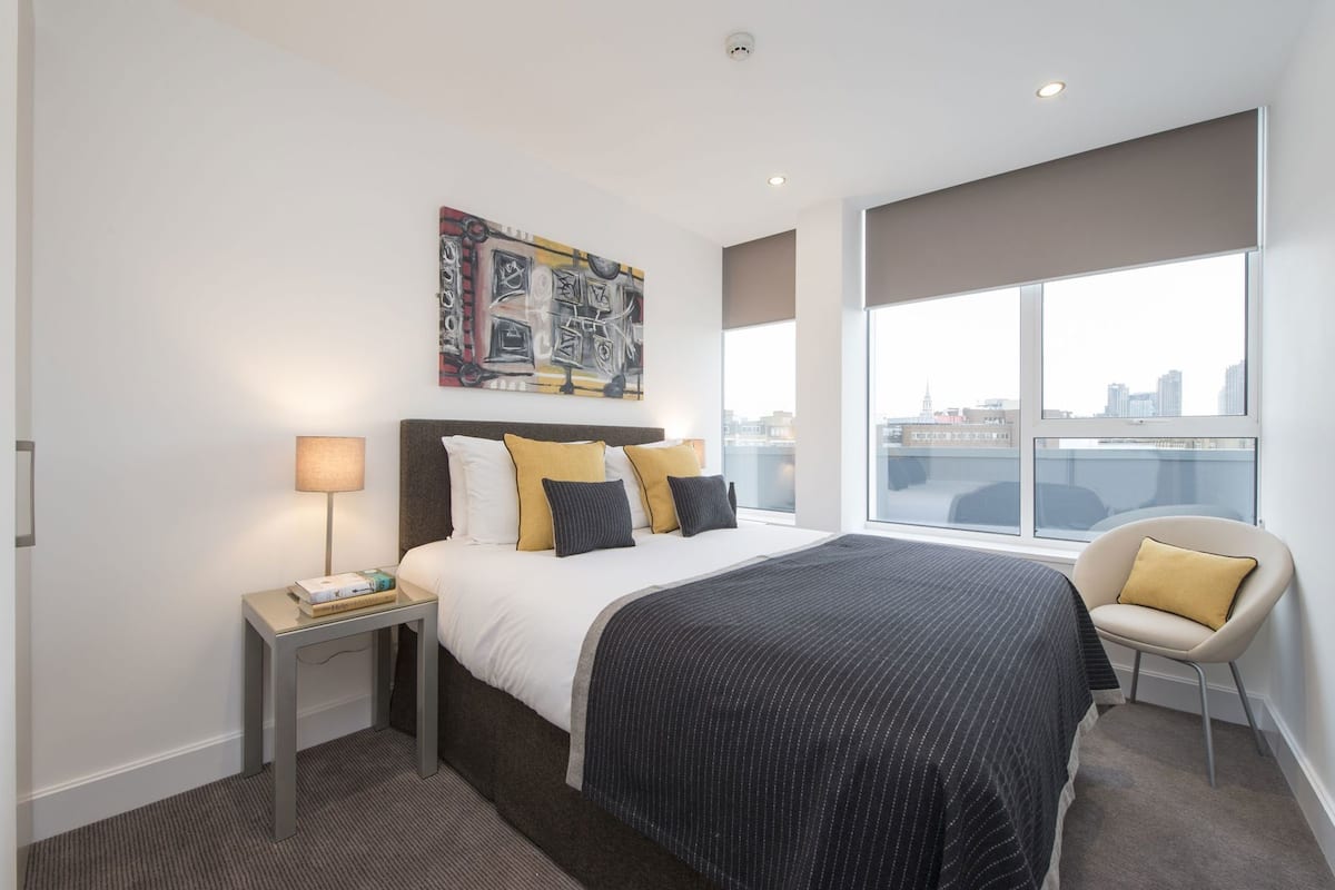 Habitación de hotel moderna con una cama grande, una mesita de noche con lámpara, una silla y vistas a la ciudad a través de amplios ventanales.
