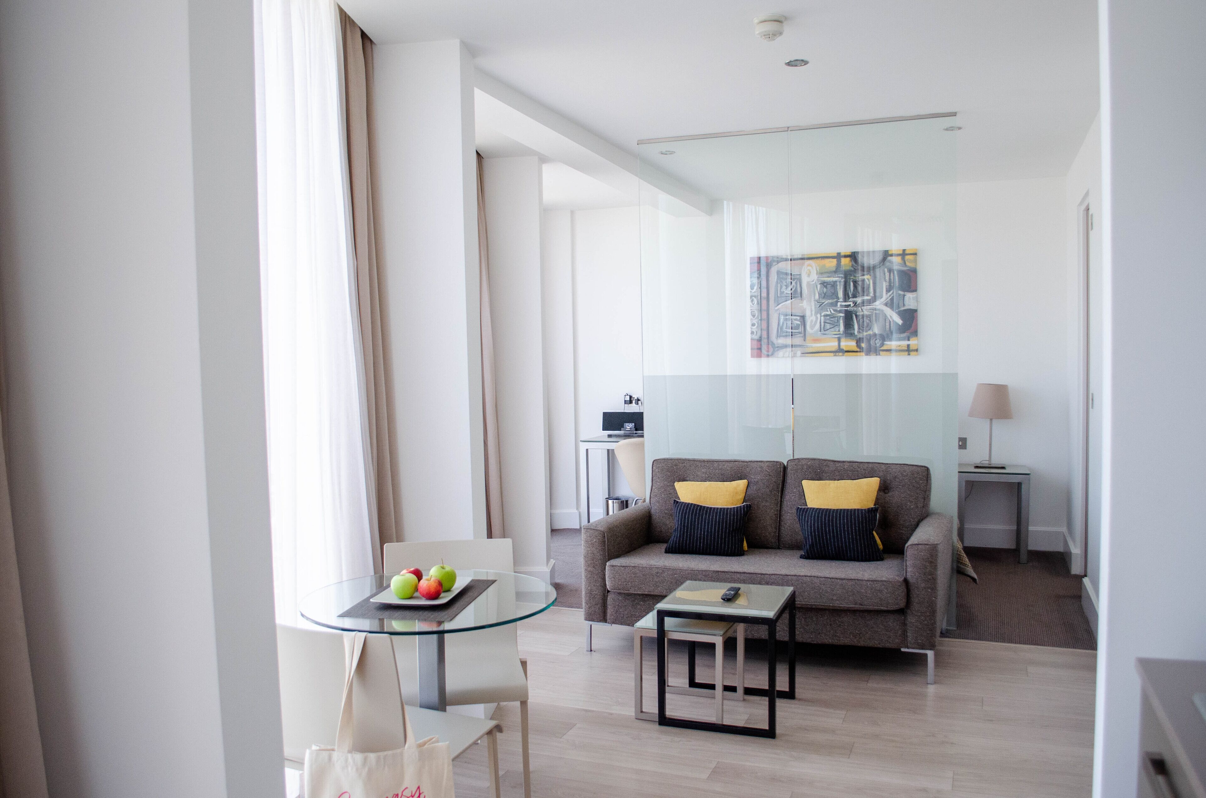 A modern living room with a glass table, a sofa with cushions, a lamp, and a painting on the wall.